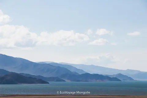A blue lake in the middle of pasture and mountains of Bayan Ölgii, the sky is light blue with some clouds