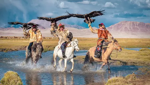 Mongolian nomads holds his golden eagle in his hand while horseriding accross a river