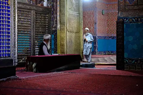 Interior of the Blue Mosque, featuring high vaulted ceilings, intricately tiled walls in shades of blue and white, and a large prayer hall carpeted in red, with soft natural light filtering through arched windows.