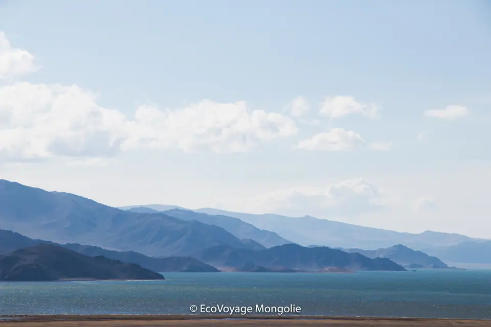 A blue lake in the middle of pasture and mountains of Bayan Ölgii, the sky is light blue with some clouds