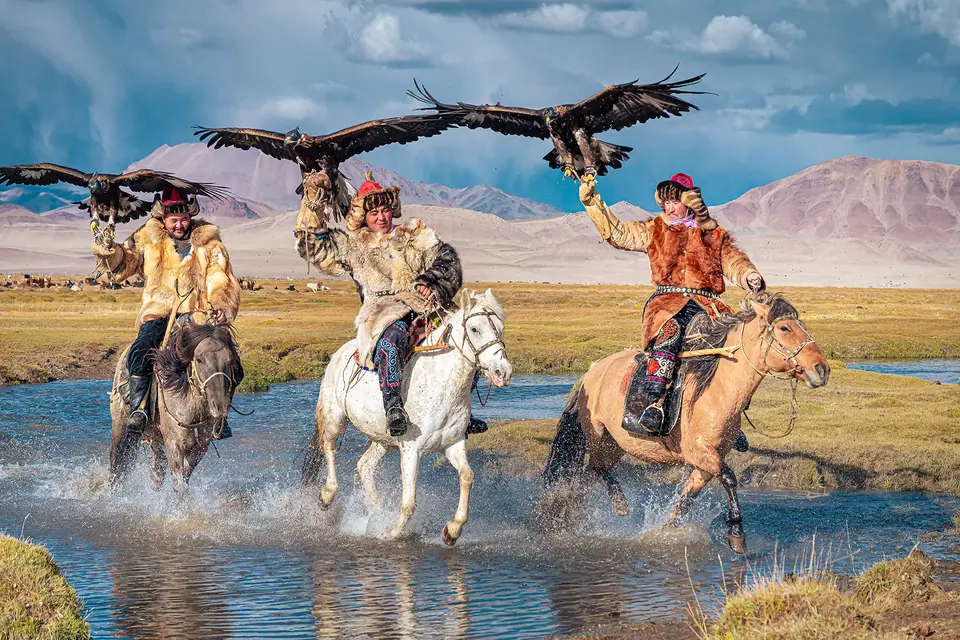 Mongolian nomads holds his golden eagle in his hand while horseriding accross a river