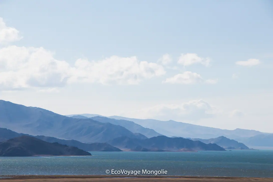 A blue lake in the middle of pasture and mountains of Bayan Ölgii, the sky is light blue with some clouds